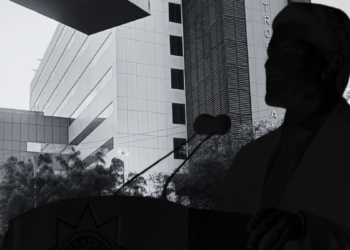 Carencias-sistema-salud-nuevo-leon - Alcaldes Regios Silhouette of a Man in a Suit Speaking at a Podium with a Microphone Against a Modern Building Backdrop. - Alcaldes Regios