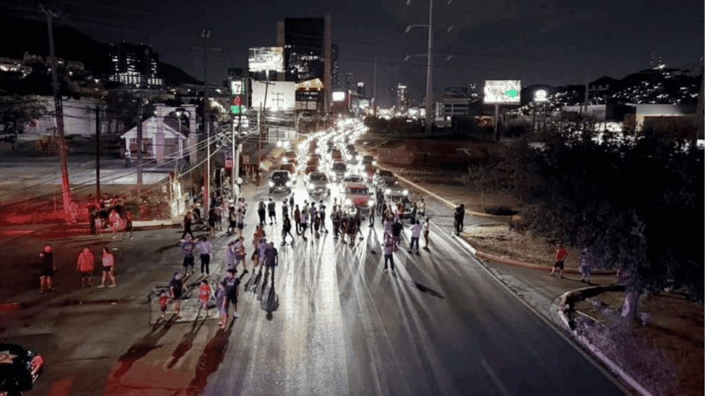 Nighttime Street Scene with a Crowd Blocking a Multi-lane Road As Cars Idle Behind Them, City Lights in the Background - Alcaldes Regios