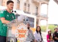 Man in a Green Jersey Speaks at a Clear Lectern During an Outdoor Event, Papers in Hand, with an Orange Bus and Spectators Behind Him. - Alcaldes Regios