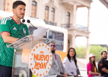 Man in a green jersey speaks at a clear lectern during an outdoor event, papers in hand, with an orange bus and spectators behind him.