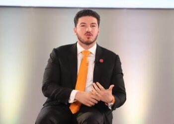 Man in a Dark Suit and Orange Tie Sits on Stage, Speaking with Hand Gestures During a Talk. - Alcaldes Regios