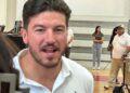 Close-up of a Man in a White Shirt Speaking to the Camera, with People and Gym Bleachers in the Background. - Alcaldes Regios