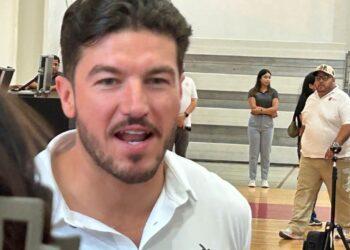 Close-up of a Man in a White Shirt Speaking to the Camera, with People and Gym Bleachers in the Background. - Alcaldes Regios